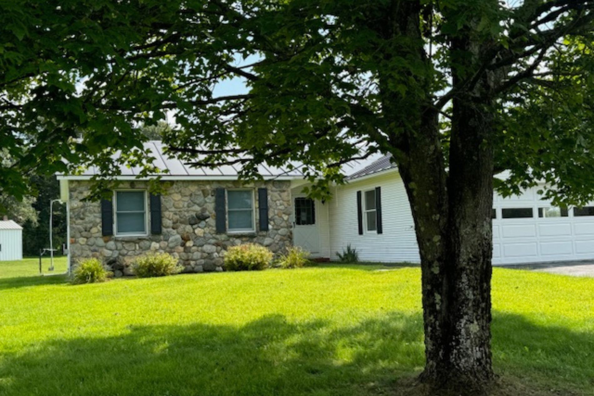 white single story house with stone on 1 wall, windows with brown shutters, large tree in yard, 2 car garage