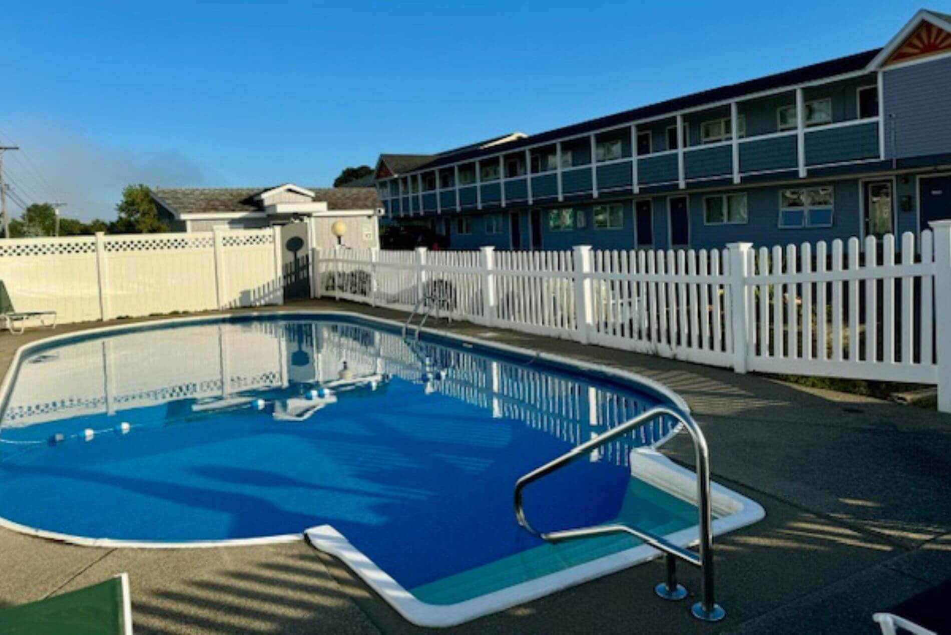 circular shaped outdoor pool with steps to walk in surrounded by fence with 2-story gray building on the right.